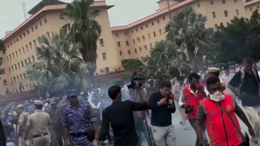 A wide street outside a government building, hazy with tear gas. A line of police and paramilitary personnel stands on the left; protesters and camera operators move away from them on the right.